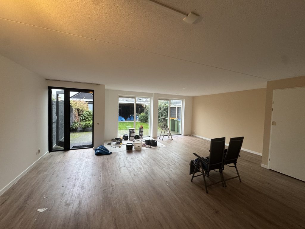 Woonkamer met tuindeuren en lichte wanden waar het behangwerk aan de gang is, met verfemmers op de vloer