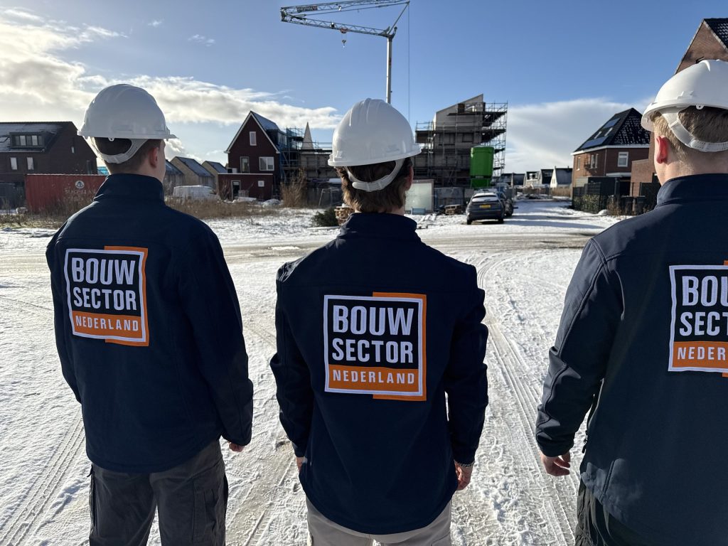 Drie vakmensen van Bouwsector Nederland met witte helm op een besneeuwde straat met huizen in aanbouw